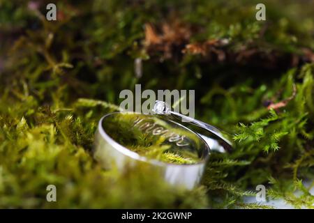 Elegante anello diamantato e anello di aggancio con iscrizione per sempre sul muschio verde. Belle luci e luce solare, messa a fuoco morbida e selettiva. Concetto di proposta di matrimonio. Foto Stock