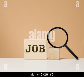 Cubetti di legno con l'iscrizione e una lente di ingrandimento nera su un tavolo bianco. Ricerca di lavoro, disoccupazione Foto Stock