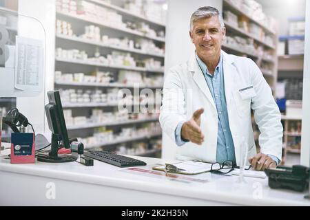 Cosa posso fare per voi oggi? Ritratto di un farmacista maschio maturo allegro che si allunga per dare una stretta di mano mentre si guarda la macchina fotografica. Foto Stock