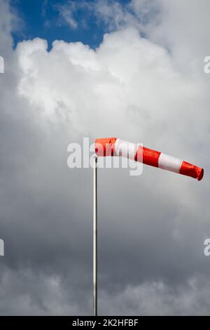 calza a vento rossa/ tubo tessile su un cielo tempestoso di giorno, di giorno, nuvole scure con il sole che si infrange Foto Stock