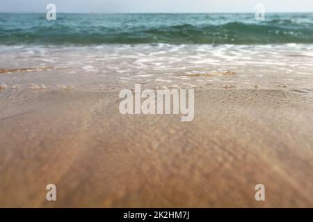 Foto a basso angolo da terra - sabbia bagnata dal mare, gocce d'acqua nell'aria, piccole onde e oceano sfocato in distanza. Sfondo astratto delle vacanze. Foto Stock