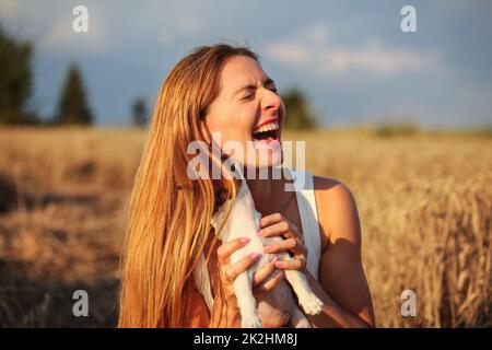 Giovane donna nel campo di grano illuminato dal sole del pomeriggio, tenendo cucciolo cercando di posa, ma cane è nascosto dietro i capelli, leccare e masticare il suo orecchio. Foto Stock
