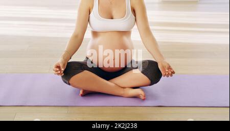 Equilibrando la sua nuova vita. Scatto corto di una giovane donna incinta irriconoscibile meditando su un tappeto yoga. Foto Stock