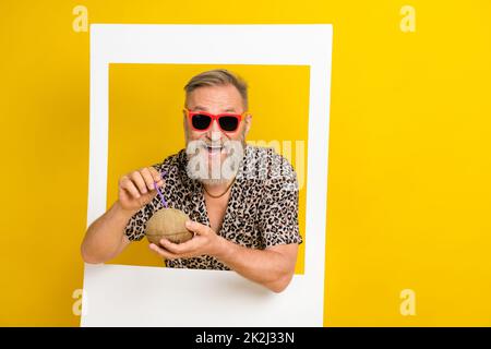 Ritratto di funky nonno braccia tenere cocktail di cocco carta album finestra di cartone isolato su sfondo giallo Foto Stock