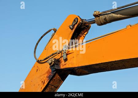 Dettaglio del braccio arancione della macchina escavatore, del meccanismo del pistone idraulico e del giunto sporco da olio nero visibile. Pomeriggio cielo blu contrasto sullo sfondo. Foto Stock
