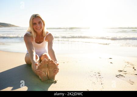 Sole, mare, sabbia è tutto ciò di cui ho bisogno. Scatto a tutta lunghezza di una giovane donna attraente in un costume da bagno che si allunga presto la mattina sulla spiaggia. Foto Stock