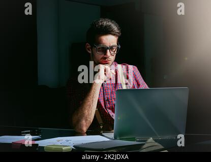 Ha messo un sacco di pensiero nei suoi disegni. Scatto corto di un bel giovane designer che lavora sul suo laptop in ufficio. Foto Stock