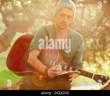 Testi ispirati dalla natura. Scatto di un bel giovane seduto in un parco con una chitarra che scrive una canzone su un blocco note. Foto Stock