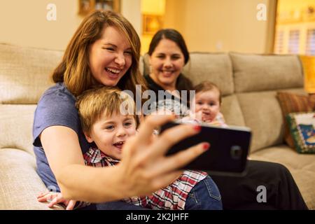 Dite che la famiglia è il nostro luogo felice. Scatto corto di due madri che prendono un selfie con i loro adorabili bambini sul divano a casa. Foto Stock