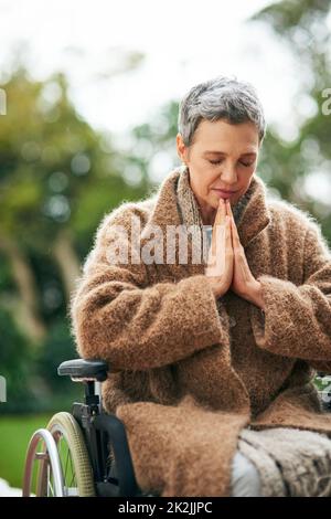 Ricerca di guida dall'alto. Scatto corto di una donna anziana che prega mentre si siede in sedia a rotelle all'aperto. Foto Stock