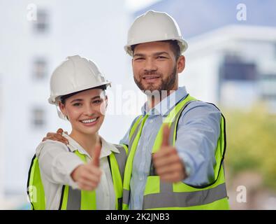 Tutto è all'altezza del codice. Ritratto ritagliato di due giovani operai edili che danno i pollici in piedi fuori dal sito. Foto Stock