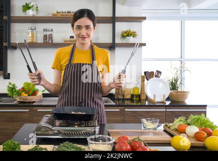 Giovane casalinga che tiene BBQ Clamp pala e forchetta in acciaio inox davanti alla pentola di cottura. Atmosfera mattutina in una cucina moderna. Il banco della cucina è pieno di vari tipi di verdure. Foto Stock