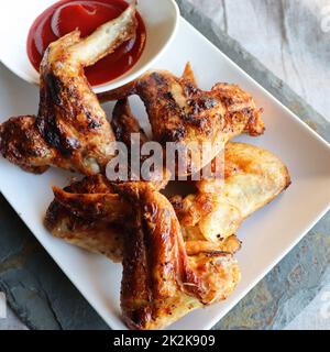 Ali di pollo speziate alla griglia con ketchup su un piatto bianco su un fondo di pietra grigia o cemento. Vista dall'alto Foto Stock