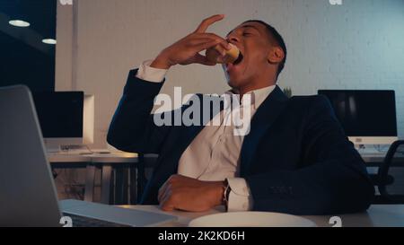 Uomo d'affari afro-americano mangiare ciambella vicino sfocato laptop in ufficio in serata, immagine di scorta Foto Stock
