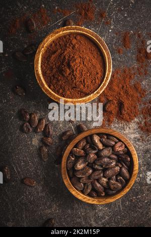 Cacao in grani e cacao in polvere in ciotole di legno su tavola nera. Foto Stock