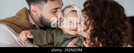 l'uomo bearded e la donna curly baciano le guance di figlia felice del bambino in fascia, bandiera, immagine di scorta Foto Stock