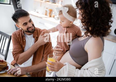 donna ricciola che tiene in braccio la figlia del bambino e che porta il succo d'arancia al marito durante la prima colazione, immagine di scorta Foto Stock