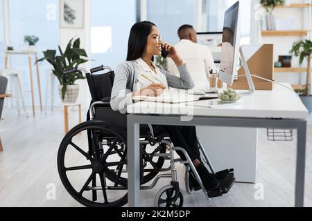 Shes sempre su chiamata e disponibile. Scatto a tutta lunghezza di una giovane donna d'affari attraente in una sedia a rotelle che fa una telefonata mentre lavora alla sua scrivania in ufficio. Foto Stock