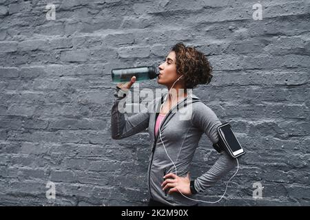 Idratare e sostituire gli elettroliti. Ripresa ritagliata di una giovane donna attraente che ascolta musica e acqua potabile dopo essersi allenata mentre fuori. Foto Stock