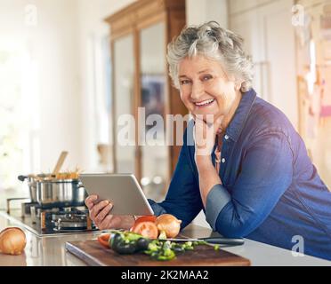 Questa ricetta viene fornita anche con un tutorial video. Ritratto di una donna anziana che usa un tablet digitale mentre cucinava in cucina. Foto Stock