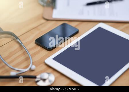 Medici essenziali quotidiani. Primo piano di un tablet, smartphone, appunti e stetoscopio su una scrivania del medico. Foto Stock