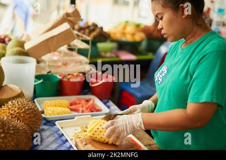 Taglio di precisione. Shot di una donna che affetta frutta in un mercato alimentare tailandese. Foto Stock