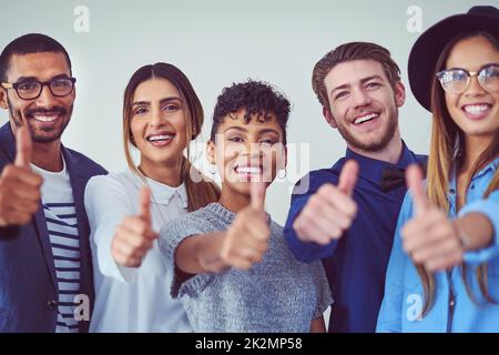 Resta al passo con il grande lavoro. Ritratto di studio di un gruppo di giovani uomini d'affari che mostrano i pollici in su mentre si alza su uno sfondo grigio. Foto Stock