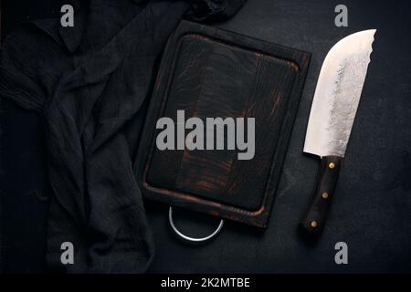 tagliere in legno vuoto rettangolare e coltello da cucina su tavola nera con tovagliolo a garza, vista dall'alto Foto Stock