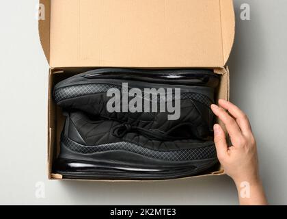 le mani femminili stanno tenendo una scatola marrone di cartone con le sneakers nere dello sport su un tavolo grigio, ispezionando un ordine in linea, vista dall'alto Foto Stock