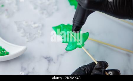 Un lecca lecca verde sotto forma di albero di Natale è tenuto nelle mani di guanti neri e bruciato sul fuoco da un accendino, nel processo di fabbricazione di un lecca. Foto Stock