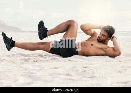 Im lavorando su quel pacchetto di sei. Scatto di un giovane sportivo che fa crunches mentre alla spiaggia per un allenamento. Foto Stock