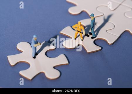 Squadra di minuscoli lavoratori in miniatura figure su jigsaw collegati puzzle pezzi isola su carta blu scuro, viola. Primo piano su elementi puzzle bianchi collegati toget Foto Stock