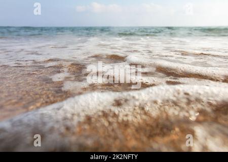Angolo basso, fotocamera a terra, obiettivo coperto da gocce d'acqua per enfatizzare l'umidità - primo piano delle onde di mare poco profonde che lavano la sabbia della spiaggia. Sfondo marino astratto. Foto Stock