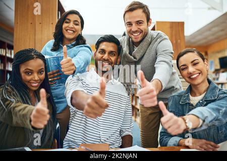 I nostri studi stanno andando grandi. Ritratto ritagliato di un gruppo di studenti universitari che ti mostrano i pollici durante lo studio in biblioteca. Foto Stock