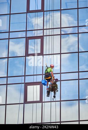 Alpinista industriale lavando finestre di vetro di un alto edificio, appeso a funi di sicurezza. Pulitori professionali per finestre che lavorano insieme al grattacielo esterno. Foto Stock