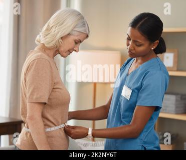 La dieta che mi hai messo mi ha fatto bene. Shot di un medico che utilizza un nastro di misurazione su uno stomaco donna anziano durante una consultazione a casa. Foto Stock