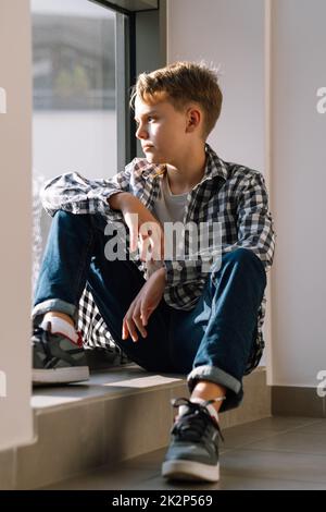 Ritratto di un adolescente carino in abiti casual. Un ragazzo giovane siede sul pavimento di un edificio e guarda fuori dalla finestra Foto Stock