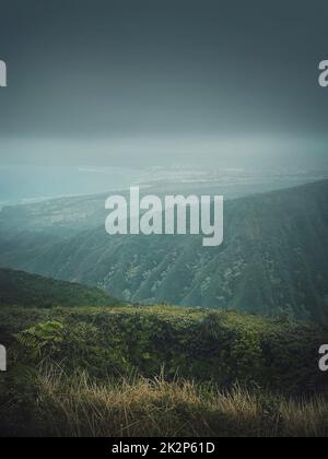 Vista panoramica dalla cima delle montagne alla costa dell'oceano alle Hawaii, isola di Oahu. Moody paesaggio escursionistico con nebbia, verdi colline all'orizzonte e nuvole tempestose sulla baia Foto Stock