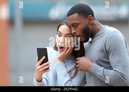 Stupito coppia interracial che controlla le notizie del telefono Foto Stock