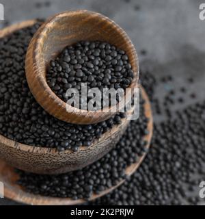 Ciotole di legno di lenticchie nere secche fagioli in primo piano sul tavolo grigio, fonte di proteine per la dieta vegetariana Foto Stock