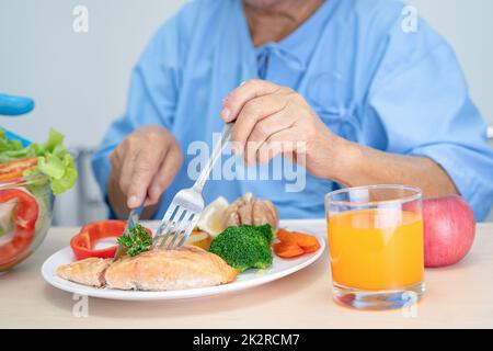 Donna anziana o anziana asiatica, paziente che mangia colazione con bistecca di salmone con cibo sano vegetale mentre si siede e affamato a letto in ospedale. Foto Stock