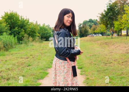 bella ragazza sorridente con un telefono sulla cintura guarda indietro mentre si trova in piedi nel mezzo di un parco verde Foto Stock