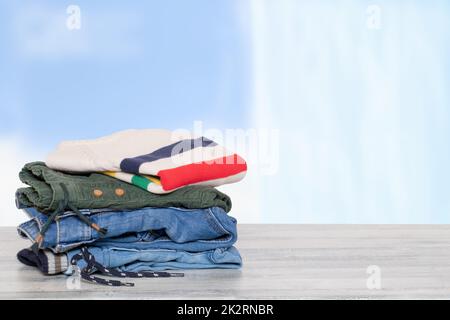 Pila di jeans. Primo piano di un mucchio di pantaloni denim colorati e di un pullover colorato su un tavolo luminoso contro astratto sfocato sfondo tenda blu chiaro. Modello per la registrazione del display del prodotto. Foto Stock