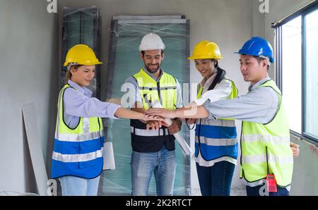 L'architetto caucasico con pianta si unisce alle mani del collega dopo aver finito l'incontro. Tutti indossano casco e giubbotto di sicurezza. Ambiente di lavoro degli ingegneri nel cantiere di progetti abitativi. Foto Stock