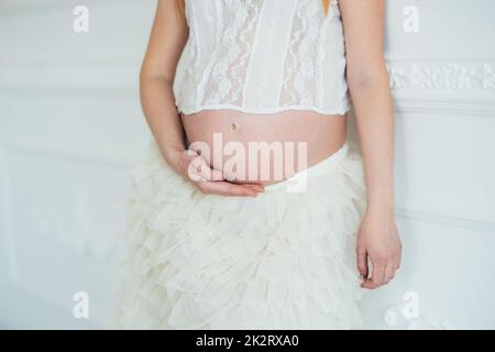 ragazza incinta dai capelli rossi in un vestito bianco vicino alla parete bianca Foto Stock