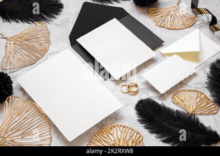 Cartoncini e busta su tavola in marmo vicino a piume nere e foglie dorate. Mockup matrimonio Foto Stock