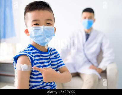 Bambino felice che indossa maschera medica e spalla con bendaggio adesivo sul braccio dopo la vaccinazione Foto Stock