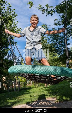 Ragazzo allegro e malizioso che dondola su un'oscillazione pubblica del parco giochi. Foto Stock