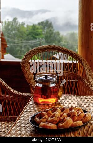 Tè di frutta calda in una teiera di vetro e biscotti si trova sul bordo di un tavolo di rattan. Foto Stock