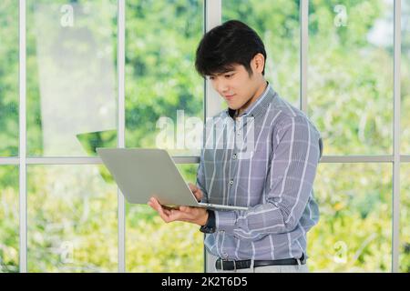 uomo d'affari sorridente e che lavora su un computer portatile stand vicino alla finestra in ufficio Foto Stock